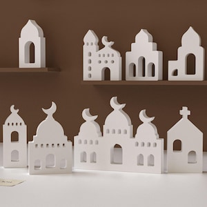 Può includere: Una collezione di modelli architettonici bianchi, tra cui moschee e una chiesa, disposti su una superficie bianca e una mensola marrone. I modelli presentano ingressi ad arco, dettagli a mezzaluna e strutture a forma di croce.