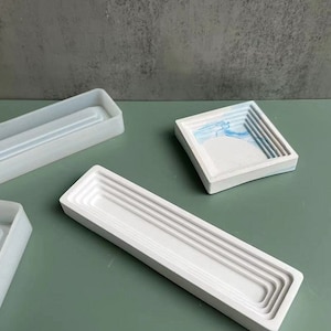 Può includere: Tre vassoi rettangolari bianchi con bordi rialzati. I vassoi sono in cemento e hanno una finitura liscia e lucida. Il vassoio a sinistra ha un design semplice, mentre gli altri due vassoi hanno un design più intricato con più anelli concentrici.