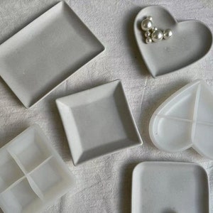 Peut inclure: Cinq plateaux en céramique de formes et de tailles différentes. Les plateaux sont tous blancs et ont une finition lisse et mate. Les plateaux sont disposés sur une surface blanche.