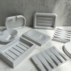 Puede incluir: Conjunto de siete jaboneras de hormigón gris con diferentes diseños, incluyendo una bandeja rectangular con nervaduras, una bandeja redonda con borde elevado y una bandeja con diseño ondulado.