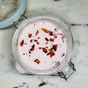 May include: A clear glass jar filled with a light pink powder and dried rose petals. The jar has a silver metal clasp and a clear glass lid. The rose petals are various shades of brown and red. The jar is sitting on a white surface.