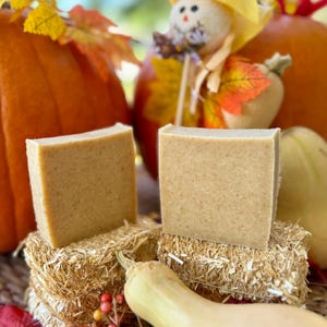 Puede incluir: Dos jabones cuadrados de color marrón claro se exhiben sobre pequeñas balas de heno, con calabazas y calabacines en el fondo. Los jabones tienen una superficie texturizada. La escena evoca un tema de cosecha de otoño.