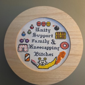 May include: A circular wooden frame containing a cross-stitch embroidery on white fabric. The text reads Unity Support Family & Kneecapping Bitches, surrounded by pixel-art style icons including skulls, potions, a treasure chest, and a banana.