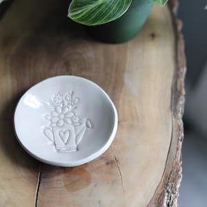 May include: A small, off-white ceramic dish with a raised floral and watering can design. The dish is round and sits on a wooden surface. A green plant in a dark green pot is in the background.