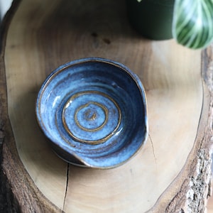 May include: A handmade blue ceramic spoon rest with a spiral design in the center. The spoon rest has a small handle and a gold-colored rim. The spoon rest is on a wooden surface.