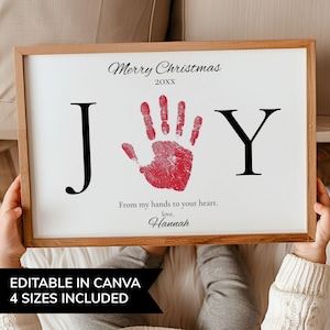 May include: A framed print with a white background and a red handprint in the center. The word "JOY" is spelled out with black letters. The text "Merry Christmas 20XX" and "From my hands to your heart, love, Hannah" are also present.