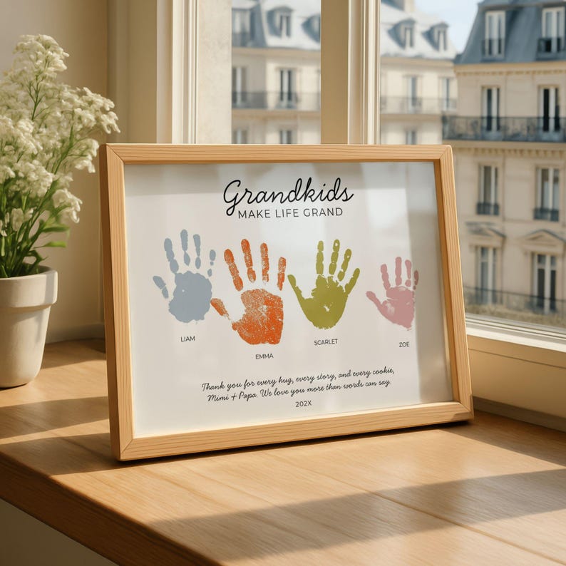 May include: A light wood framed print featuring the text Grandkids MAKE LIFE GRAND, four colourful handprints in blue, orange, green, and pink labelled Liam, Emma, Scarlet, and Zoe, and the text Thank you for every hug, every story, and every cookie, Mimi + Papa. We love you more than words can say 202X.