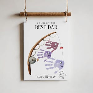 K&ouml;nnte beinhalten: Ein h&auml;ngender Wanddruck mit einer Angelruten-Illustration und drei Handabdruck-Fischen in Violettt&ouml;nen. Der Text lautet WE CAUGHT THE BEST DAD, 20XX, HAPPY BIRTHDAY und Love, Emma, Cameron & Lila. Der Druck ist an einem Holzstab befestigt.