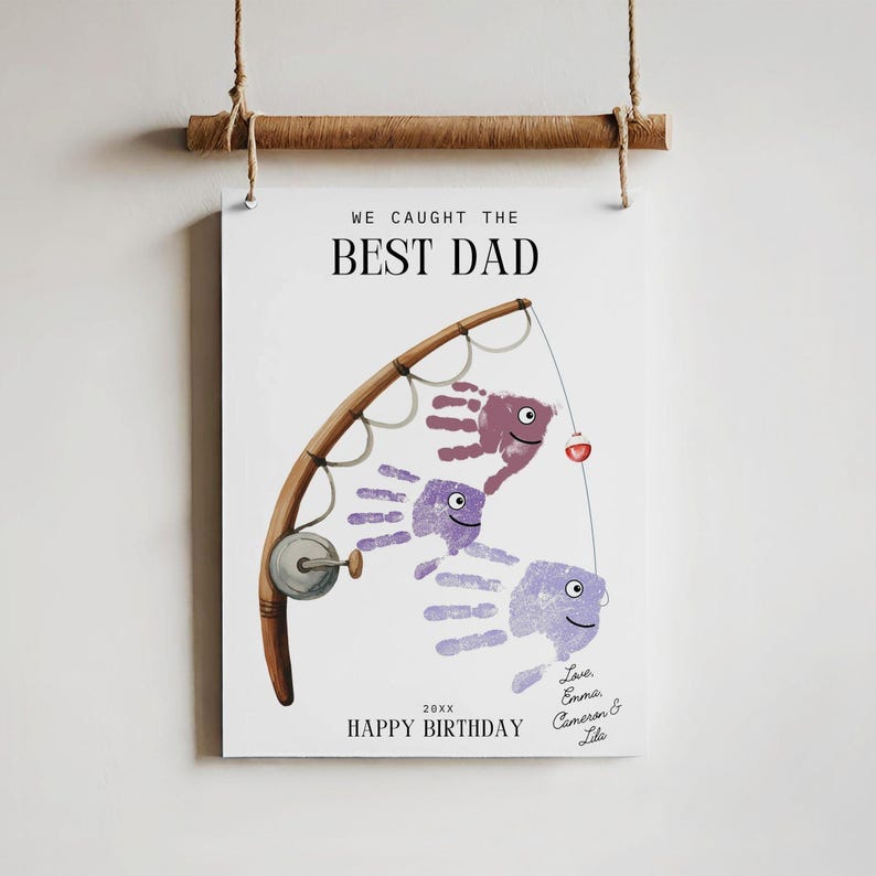 K&ouml;nnte beinhalten: Ein h&auml;ngender Wanddruck mit einer Angelruten-Illustration und drei Handabdruck-Fischen in Violettt&ouml;nen. Der Text lautet WE CAUGHT THE BEST DAD, 20XX, HAPPY BIRTHDAY und Love, Emma, Cameron & Lila. Der Druck ist an einem Holzstab befestigt.