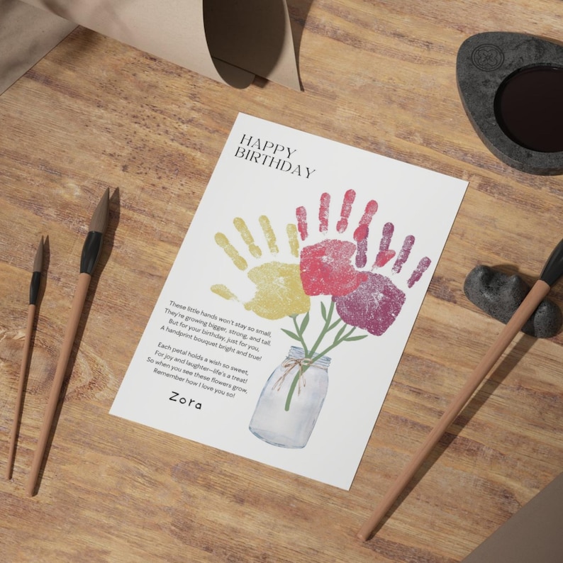 May include: A white rectangular card featuring the text Happy Birthday and a poem above a handprint bouquet illustration. The bouquet consists of yellow, red, and purple handprints resembling flowers in a glass jar. The name Zora is printed below.