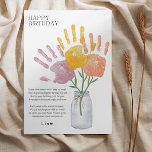 May include: A white birthday card featuring the text HAPPY BIRTHDAY and a poem about handprints. The design shows a clear glass mason jar with green stems topped by three colorful handprint flowers in yellow, orange, and purple. The name Liam is printed at the bottom.
