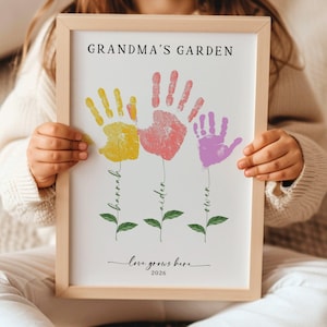Puede incluir: Impresión enmarcada con el texto "Grandma's Garden" y flores de huellas de manos en amarillo, rosa y morado. Los nombres "Barrah", "Aiden" y "Owen" están escritos debajo de cada huella. La frase "love grows here" y el año 2026 también están incluidas.