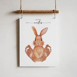 Könnte beinhalten: Eine weiße Karte mit dem Text "Frohe Ostern" und einem braunen Hasen mit braunen Fußabdrücken.