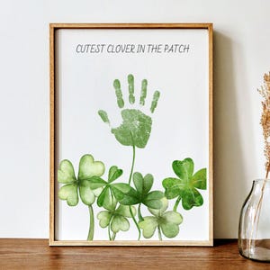 Puede incluir: Impresión enmarcada con una huella de mano verde y tréboles de acuarela. El texto "CUTEST CLOVER IN THE PATCH" está en la parte superior. La obra de arte está en un marco de madera. Un jarrón con una planta seca está en el fondo.
