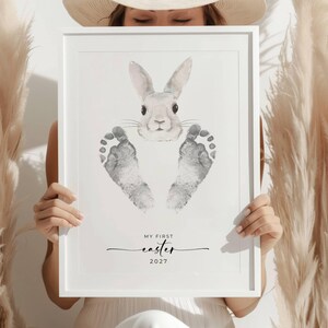 May include: A framed print with a watercolour illustration of a rabbit's head and two baby footprints. The words "MY FIRST EASTER 2027" are written in black script. The frame is white.