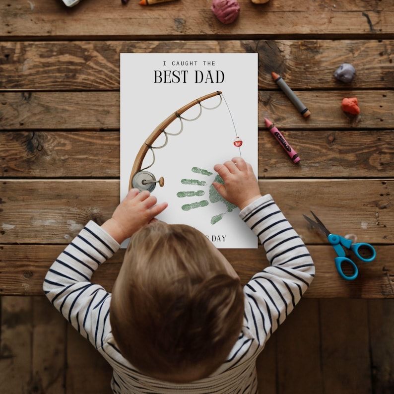 May include: A white rectangular paper featuring the black text I CAUGHT THE BEST DAD and a brown fishing rod illustration. A green handprint is positioned as the fish, with a red and white bobber attached to the line. The paper includes the text S DAY.