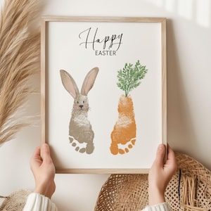 Puede incluir: Una impresión enmarcada con las palabras "Happy Easter" en escritura negra. La obra de arte presenta un conejito y una zanahoria, ambos creados a partir de huellas de pies. El conejito es gris y la zanahoria es naranja, con hojas verdes.