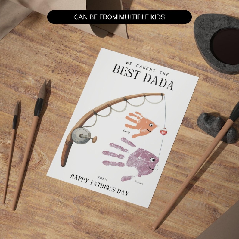 May include: A white rectangular card featuring the text WE CAUGHT THE BEST DADA and HAPPY FATHER'S DAY. The design includes a brown fishing rod illustration and two handprints in orange and purple, styled as fish with small eyes and fins.
