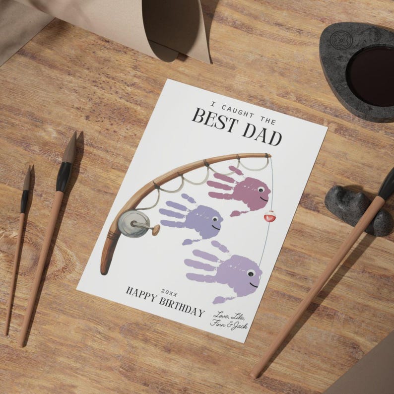 K&ouml;nnte beinhalten: Eine wei&szlig;e, rechteckige Gru&szlig;karte mit einer braunen Angelruten-Illustration und drei violetten Handabdruck-Fischen an der Schnur. Der Text lautet I CAUGHT THE BEST DAD, 20xx, HAPPY BIRTHDAY sowie Love, Lilia, Finn & Jack in schwarzer Schrift.