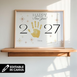 May include: A framed print with the text "HAPPY New Year!" and the year "2027". The design includes a gold handprint, gold starbursts, and the name "Olivia". The frame is light brown and sits on a wooden shelf.