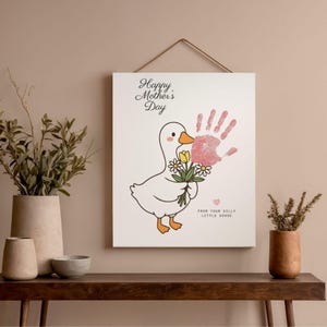 May include: A white canvas print with a cartoon goose holding flowers and a pink handprint. The text "Happy Mother's Day" and "From Your Silly Little Goose" are written on the canvas. The print is hanging on a wall above a wooden table.