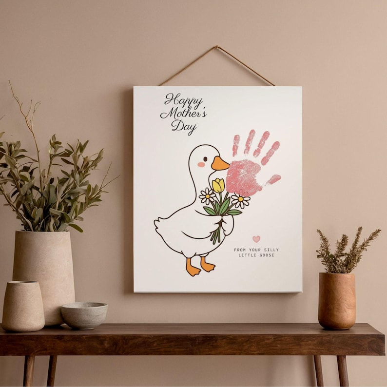 May include: A white canvas print with a cartoon goose holding flowers and a pink handprint. The text "Happy Mother's Day" and "From Your Silly Little Goose" are written on the canvas. The print is hanging on a wall above a wooden table.
