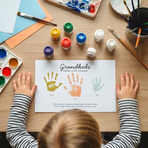 May include: A white rectangular paper print featuring the text Grandkids MAKE LIFE GRAND, three handprints in yellow, orange, and light green with the names Ethan, Lou, and Jack, and a printed message, surrounded by paint supplies on a wooden surface.