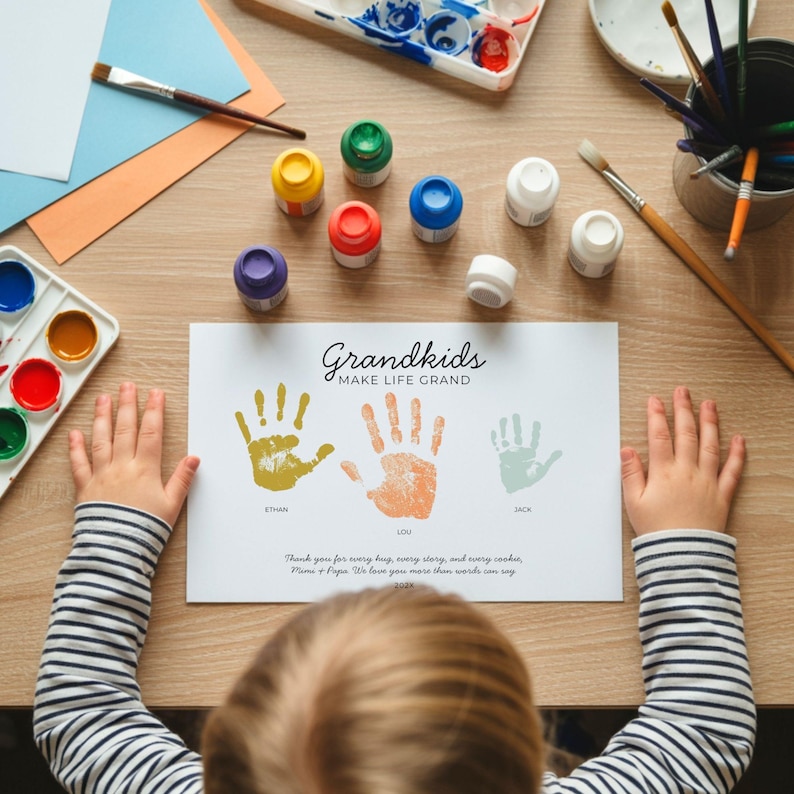 May include: A white rectangular paper print featuring the text Grandkids MAKE LIFE GRAND, three handprints in yellow, orange, and light green with the names Ethan, Lou, and Jack, and a printed message, surrounded by paint supplies on a wooden surface.