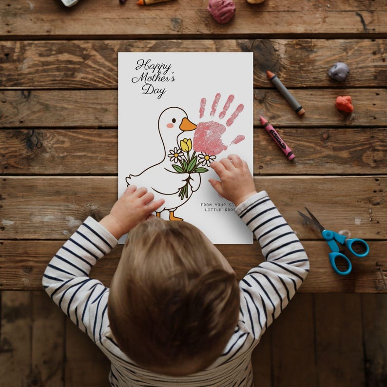 May include: A child's handmade Mother's Day card featuring a cartoon duck holding flowers and a pink handprint. The card reads "Happy Mother's Day" and "From Your Little Goose". The child is wearing a striped shirt.