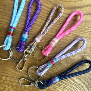 Könnte beinhalten: Fünf Schlüsselanhänger aus geflochtener Kordel in Hellblau, Lila, Bunt, Pink und Dunkelblau. Jeder Anhänger verfügt über einen Metallring und einen Karabinerhaken sowie einen dekorativen gewickelten Bereich in der Nähe der Metallteile.