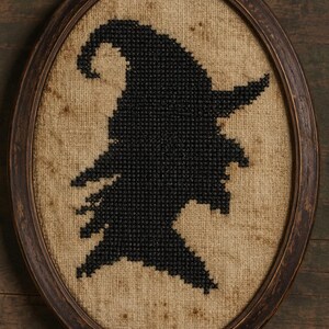 May include: An oval-shaped wooden frame containing a cross-stitch embroidery on a tan, textured fabric background. The embroidery depicts a solid black silhouette of a pointed hat with a curved top, centered within the frame.