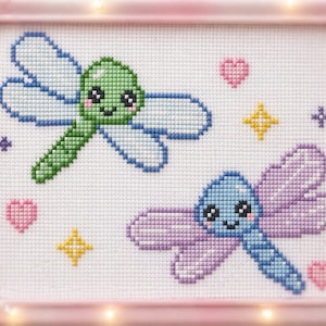 Può includere: Ricamo a punto croce con due libellule sorridenti con corpi e ali blu e verdi. L'opera d'arte include cuori rosa, stelle gialle e accenti viola, il tutto su uno sfondo bianco all'interno di una cornice rosa.
