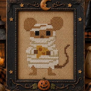 May include: A cross-stitch embroidery of a mouse dressed as a mummy, holding a yellow candy corn. The design is on tan fabric, framed in a black wooden frame decorated with small stars, a crescent moon, and a carved pumpkin at the bottom.