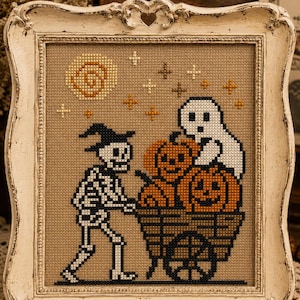 Pode incluir: Obra de arte em ponto cruz em uma moldura branca desgastada. A imagem apresenta uma cena de Halloween com um esqueleto empurrando um carrinho cheio de abóboras, um fantasma, uma lua e estrelas. A obra é bordada em tecido bege.