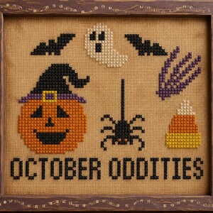 May include: A framed cross-stitch sampler on tan fabric featuring Halloween motifs including a jack-o'-lantern wearing a witch hat, a ghost, two black bats, a purple skeletal hand, a spider, and candy corn, with the text OCTOBER ODDITIES.