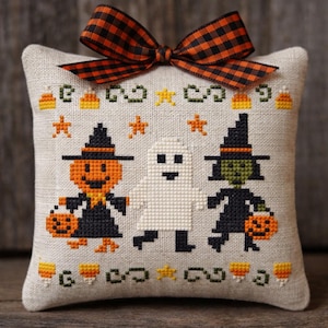 May include: A cross-stitch Halloween cushion featuring a pumpkin, a ghost, and a witch, each with a trick-or-treat bag. The cushion is decorated with candy corn and star motifs, and a plaid orange and black bow.