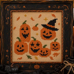 May include: A cross-stitch embroidery featuring five orange jack-o'-lanterns with black facial features, one wearing a black witch hat, set against a tan fabric background. The piece is enclosed in a square, distressed black wooden frame.