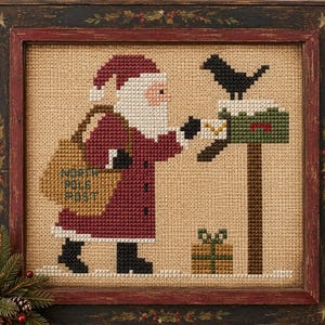 Op de afbeelding: Een kruissteekborduurwerk in een donkere houten lijst met de Kerstman in een rode jas en muts, met een beige tas waarop NORTH POLE POST staat. De Kerstman stopt een brief in een groene brievenbus met sneeuw en een zwarte vogel erbovenop.