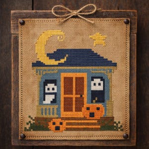 Pode incluir: Cena de Halloween em ponto cruz com uma casa azul com telhado azul escuro, uma lua crescente amarela e uma estrela amarela. A casa tem uma porta castanha e abóboras laranja. A obra de arte é emoldurada em uma moldura de madeira com um laço de barbante.