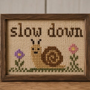 May include: A framed cross-stitch embroidery on beige fabric featuring a brown snail in the centre, flanked by a purple flower on the left and a pink flower on the right, with the text "slow down" stitched in dark brown letters above the snail.