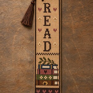 May include: A rectangular cross-stitch bookmark on tan fabric featuring the word READ in dark brown block letters, small heart and flower motifs, and a stack of four colorful books at the bottom, finished with a dark brown thread tassel.