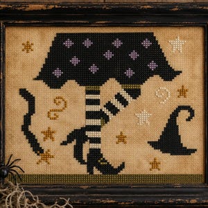 May include: A framed cross-stitch embroidery on tan fabric featuring a black skirt with purple diamond patterns, black and white striped legs, and black pointed shoes. The design includes small gold and white stars, a black hat, and a black snake.