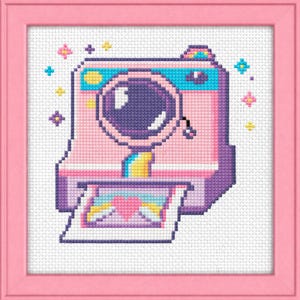 May include: A cross-stitch framed art piece featuring a pink and purple camera with a large lens. The camera is emitting sparkles and a photo is emerging with a rainbow and heart design. The frame is pink.