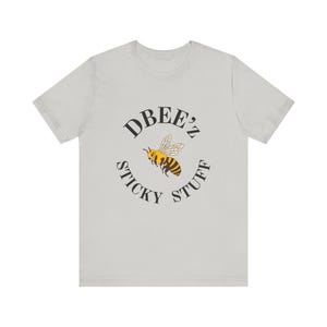 Pode incluir: Camiseta cinza claro com um design circular. O design inclui as palavras "DBEE'Z STICKY STUFF" e uma abelha de desenho animado. A abelha é amarela e preta com asas brancas. A camiseta é feita de material macio.
