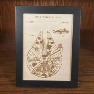 May include: A black rectangular frame containing a light-colored wooden panel with a laser-engraved technical drawing of the Millennium Falcon. The print features the text Millennium Falcon, No. 123,456, and Apr 15, 1977, along with Fig 1 and Fig 2 labels.