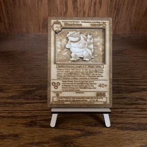 May include: A wooden laser-cut Pokémon trading card featuring the character Blastoise. The card is displayed on a small wooden easel. The card has text and a graphic of Blastoise.