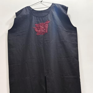 May include: Black sleeveless tunic with a round neckline and a front slit. The tunic features a textured, burgundy-colored appliqué on the chest. The garment is hanging on a hanger against a white background.