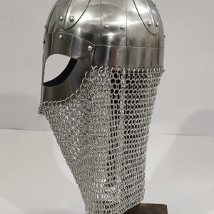 May include: A silver Viking helmet with a chainmail face mask. The helmet is on a wooden stand.