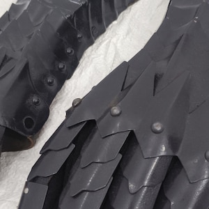 May include: A pair of black metal gauntlets with a scaly design. The gauntlets are made of metal plates that are riveted together. The gauntlets are designed to protect the hands and fingers during combat.