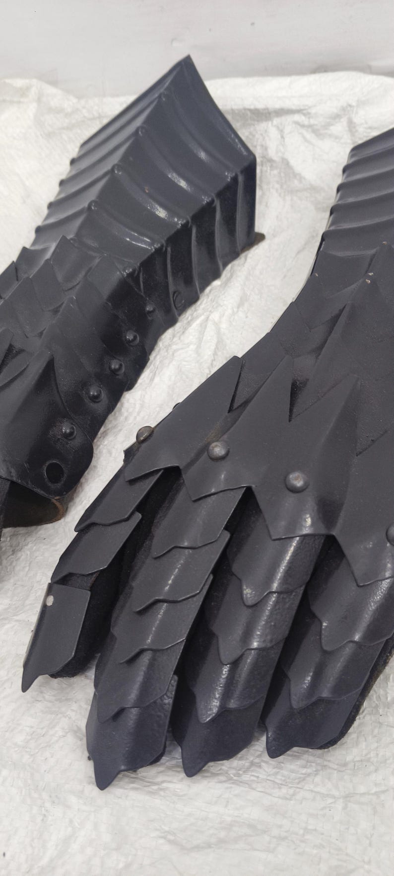 May include: A pair of black metal gauntlets with a scaly design. The gauntlets are made of metal plates that are riveted together. The gauntlets are designed to protect the hands and fingers during combat.